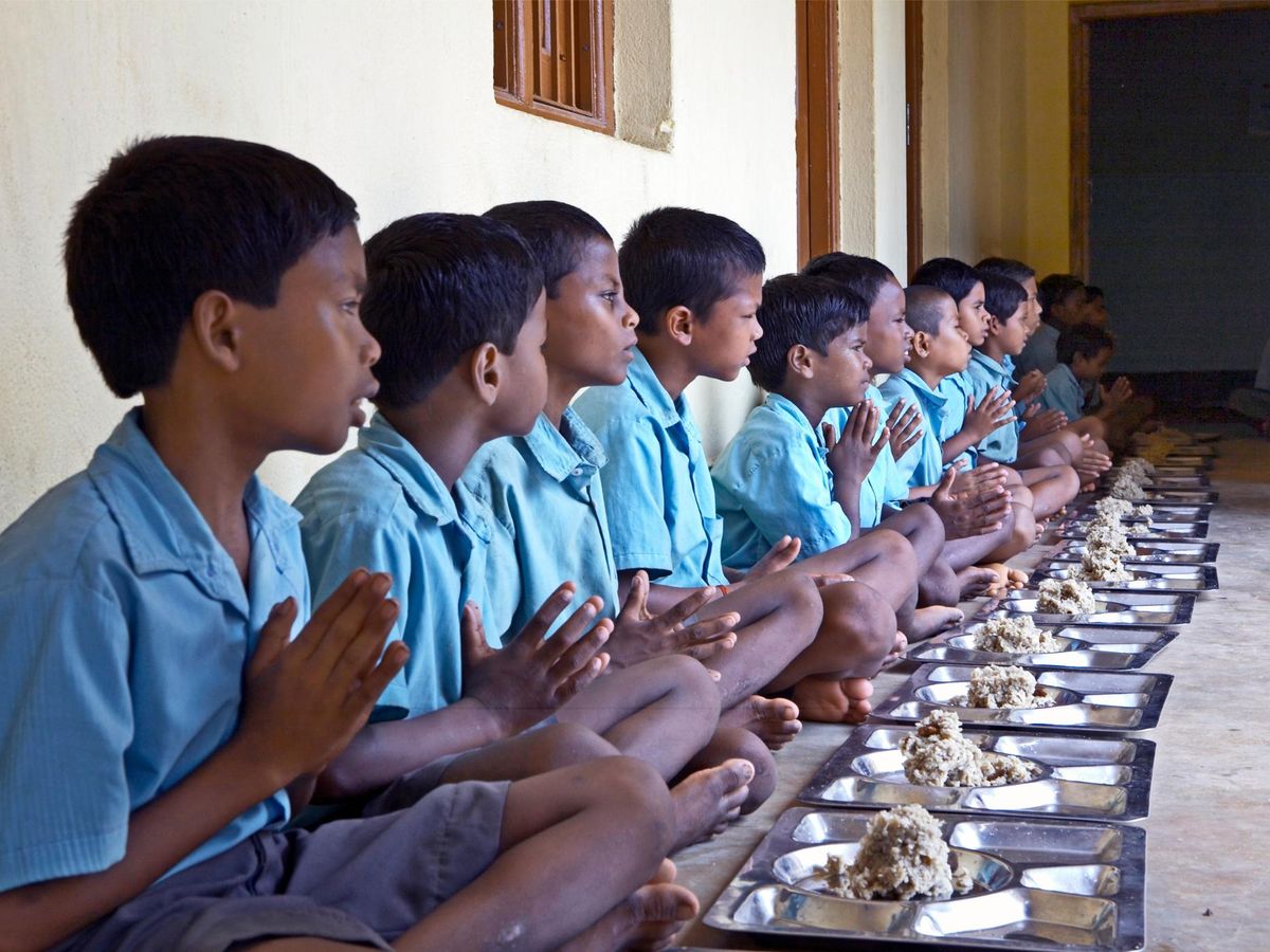 A long row of children sitting on the floor with their hands together in front of plates of food.